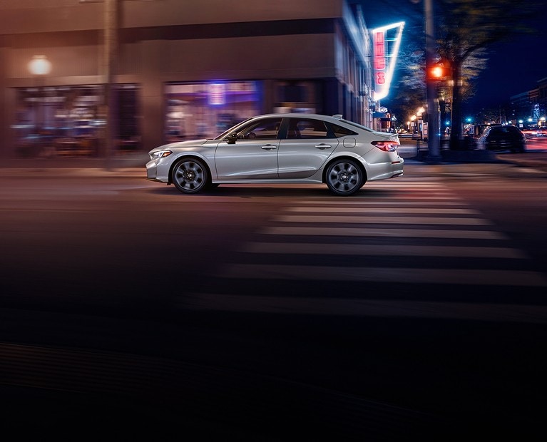 2025 Honda Civic Sedan Sport Touring Hybrid in Solar Silver Metallic driving on urban street at night - Opens a dialog 2025 Honda Civic Sedan Sport Touring Hybrid in Solar Silver Metallic driving on urban street at night - Opens a dialog